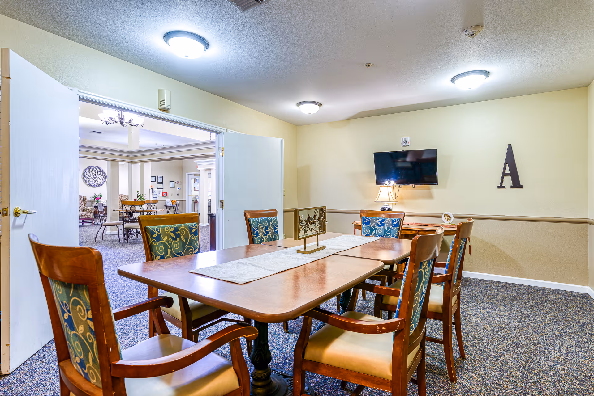 A well-lit room with a wooden table surrounded by six chairs with patterned cushions. A flat-screen TV is mounted on the wall above a small wooden cabinet with a lamp. The walls are painted beige with a darker beige wainscoting. Double doors open to an adjacent room with additional seating and decorative elements.