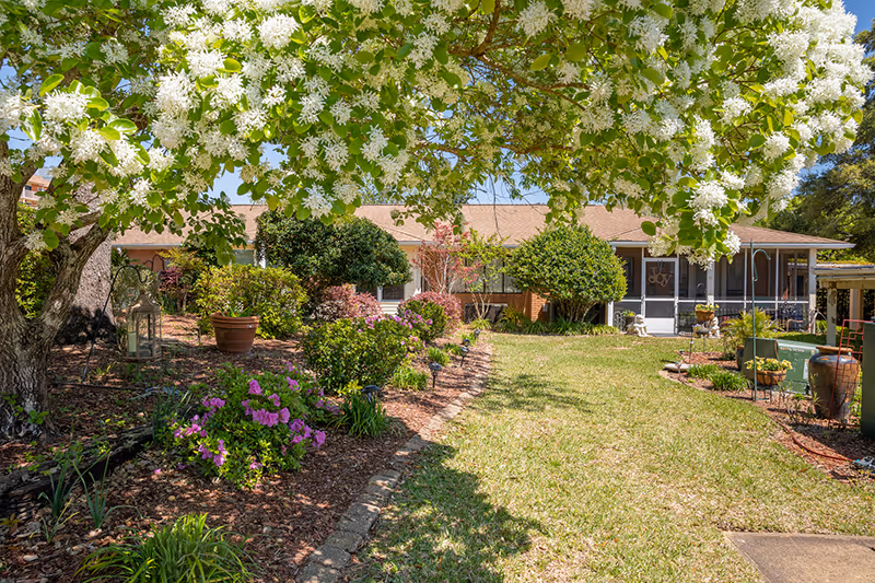 A well-maintained garden area with blooming white flowers on a tree in the foreground, colorful shrubs, and a grassy lawn leading to a single-story building with a screened porch under a clear blue sky.