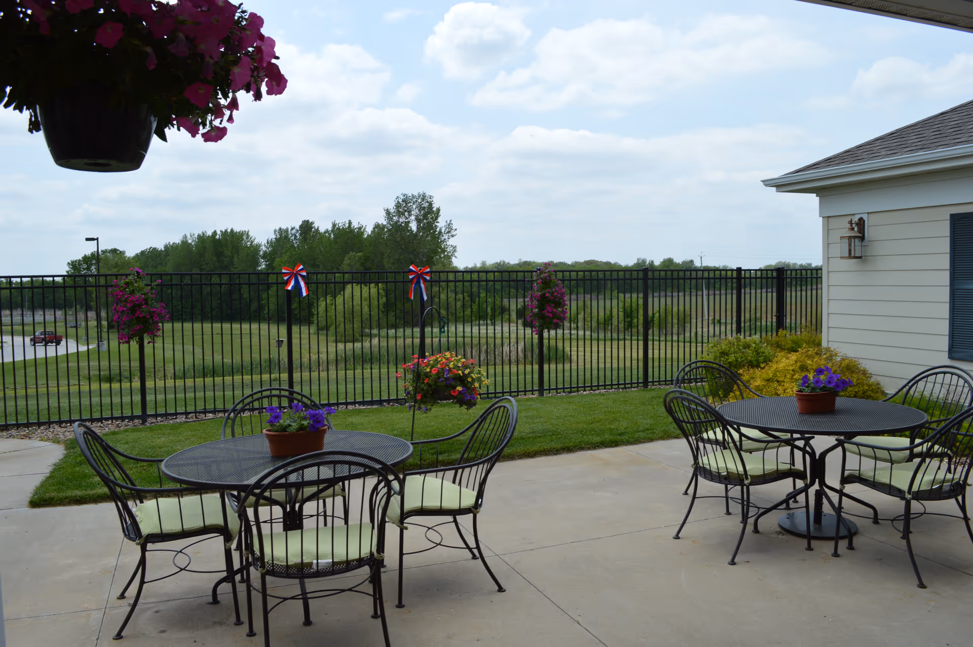 Outdoor patio area with two round metal tables and several metal chairs with light green cushions. Each table has a small potted plant with purple flowers. Hanging flower baskets and red, white, and blue ribbon decorations are attached to a black metal fence. In the background, there is a grassy field and trees under a partly cloudy sky.