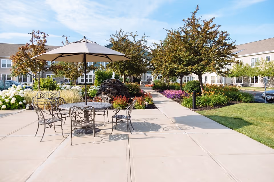 Outdoor patio area at Eddy Hawthorne Ridge with a round metal table and six matching chairs under a large beige umbrella. The patio is surrounded by well-maintained landscaping including green grass, flowering plants, bushes, and trees. In the background, there is a building with multiple windows and a walkway leading to the entrance.