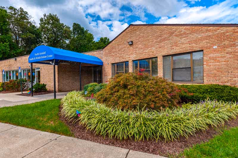 Exterior view of a single-story brick building with a blue awning labeled 'North Arundel Health & Rehabilitation Center'. The building is surrounded by green shrubs and plants, with a sidewalk leading to the entrance. The sky is partly cloudy.