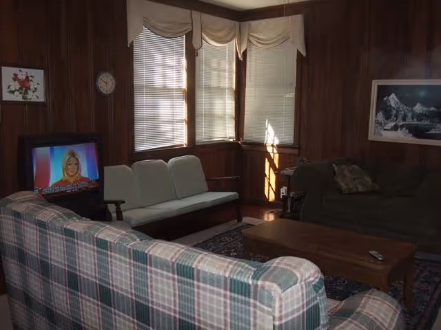 A cozy living room with wood-paneled walls, three windows with blinds and valances, a plaid sofa in the foreground, a green sofa with pillows, a wooden coffee table with a remote control, a small TV showing a news anchor, a clock on the wall, and framed artwork.