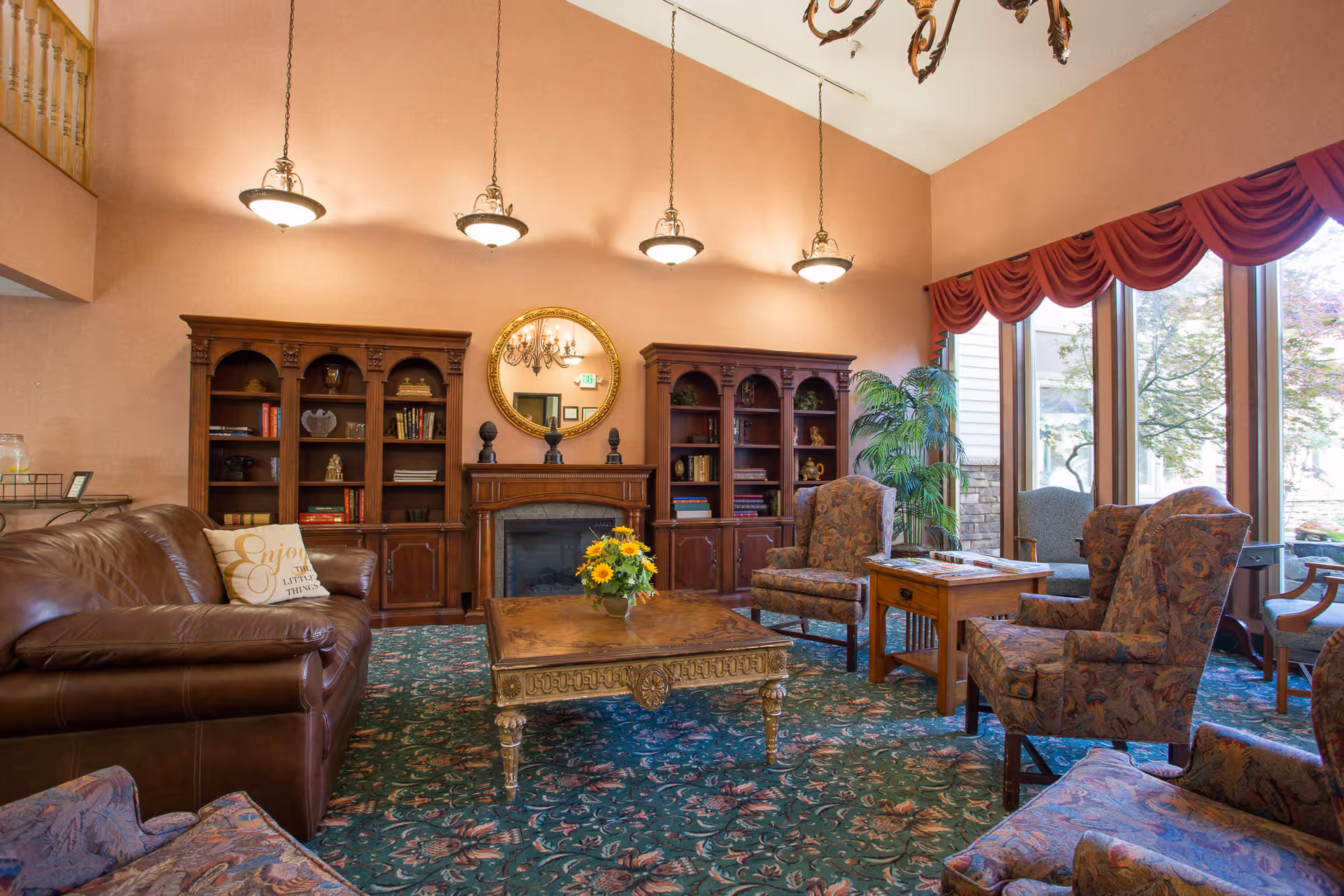 A cozy living room in Cascade Park Retirement Community featuring a brown leather sofa with a decorative pillow, several upholstered armchairs, a wooden coffee table with a flower arrangement, two wooden bookshelves flanking a fireplace with a round mirror above it, large windows with red valances letting in natural light, and a green patterned carpet.