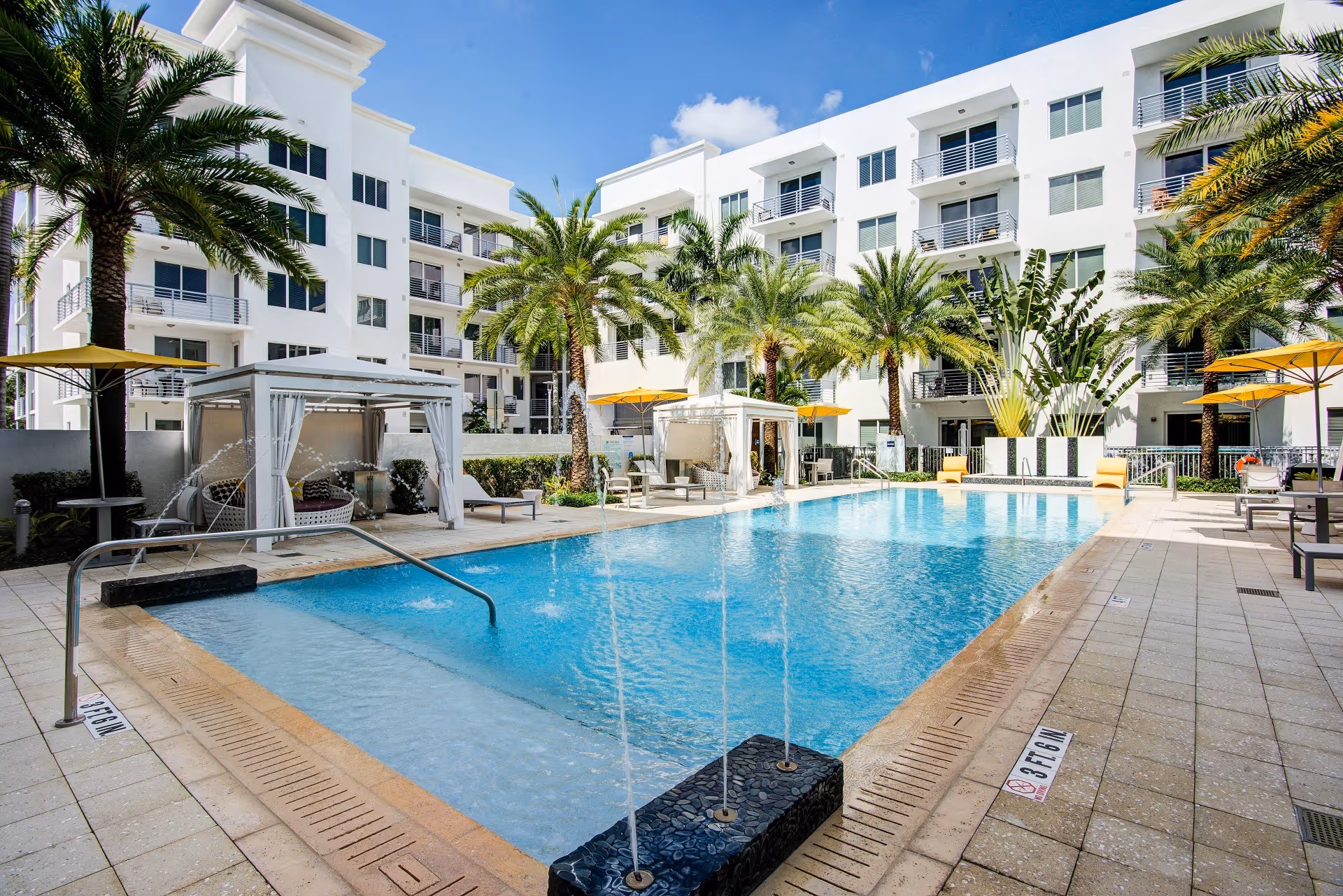 Outdoor swimming pool area at UpsideHoM Pembroke Pines with clear blue water, palm trees, white cabanas with curtains, yellow umbrellas, lounge chairs, and a modern white multi-story building in the background under a blue sky with some clouds.