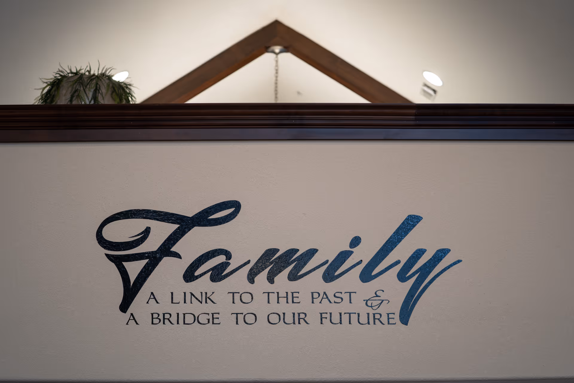 Interior wall sign reading "Family" with the quote "A link to the past & a bridge to our future" beneath a railing and plant above.