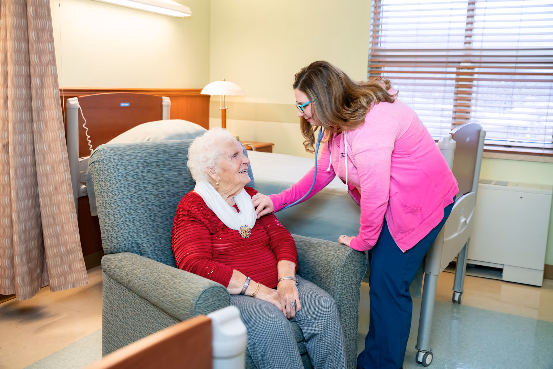 A healthcare worker in a pink jacket uses a stethoscope to check the heartbeat of an elderly woman sitting in a green armchair in a nursing facility room with a hospital bed and window blinds in the background.