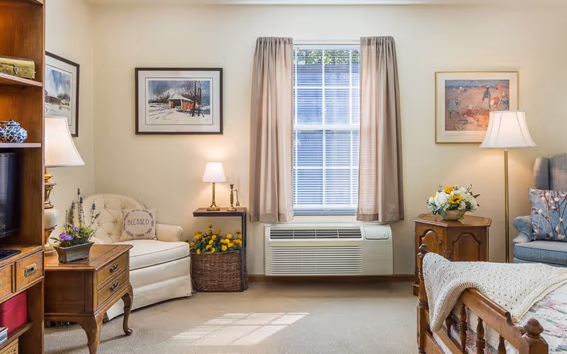 A cozy, well-lit living room with a window in the center covered by beige curtains. To the left, there is a white armchair with a pillow that says 'BLESSED' next to a small wooden side table with a lamp and a basket of yellow flowers underneath. On the right side, there is a wooden nightstand with a flower arrangement and a floor lamp beside a blue upholstered chair with patterned pillows. The room has framed artwork on the walls and a wooden bed frame partially visible on the right.