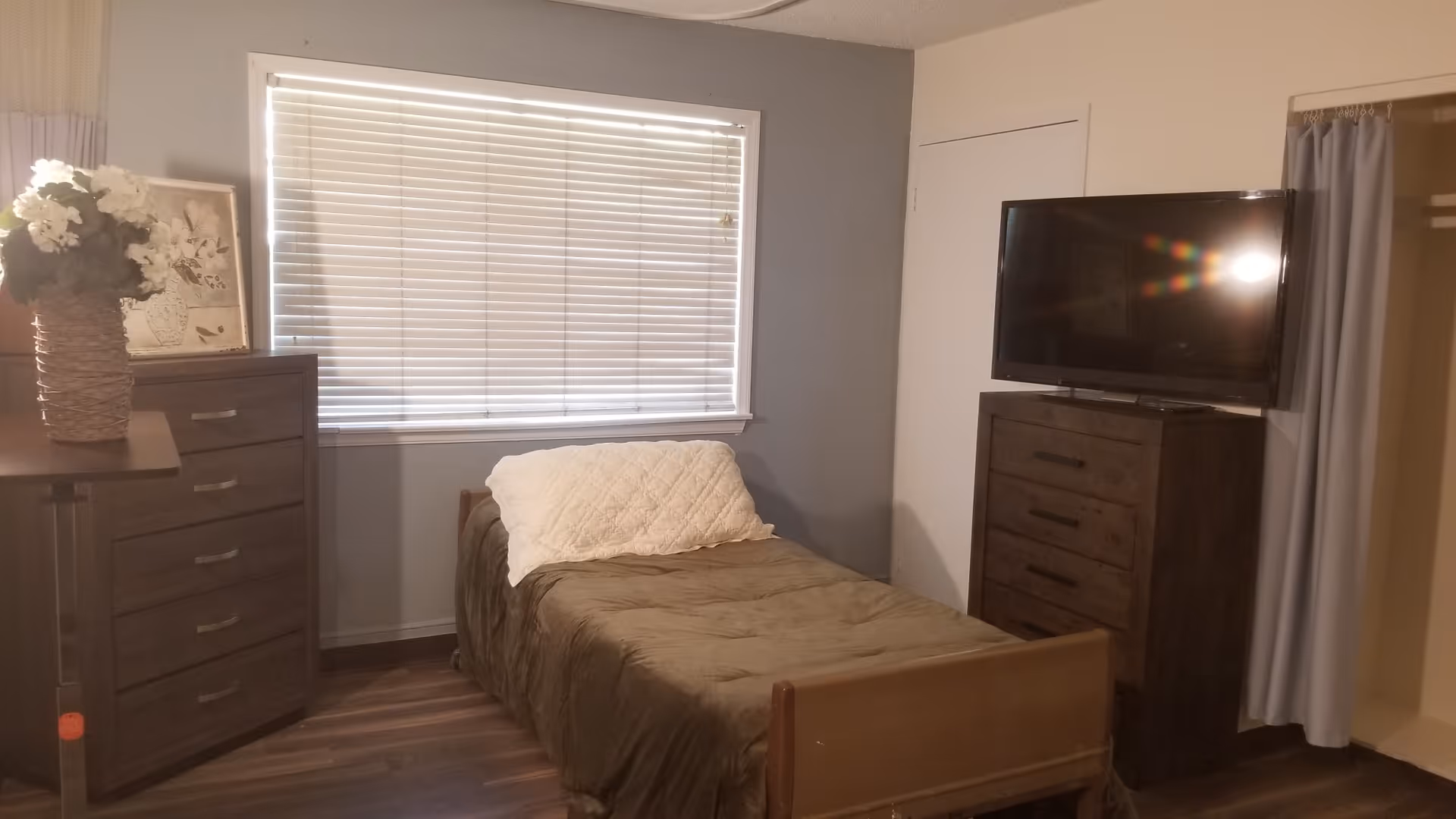 A small bedroom with a single bed covered in a brown blanket and a white pillow. There is a window with closed blinds behind the bed. To the left of the bed is a dark wooden dresser with a vase of flowers and a framed picture on top. To the right of the bed is another dark wooden dresser with a flat-screen TV on top. A closet with a curtain is partially visible on the right side of the room. The floor is wood, and the walls are painted light gray.