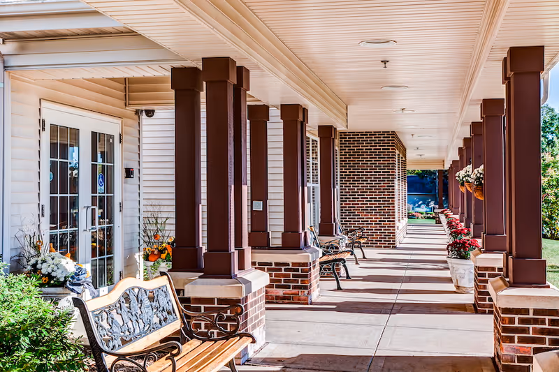 Covered outdoor walkway at a senior living facility with brick columns and wooden benches along the sides. Flower pots with blooming flowers are placed on the ground and hanging from the ceiling. Double glass doors lead into the building on the left side.