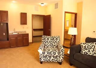 Interior view of a senior living facility room with beige walls and carpet. The room features a patterned armchair, a dark sofa with matching patterned pillows, a wooden side table with a lamp, a small kitchenette with a sink and cabinets, and an open door leading to a bathroom with a visible sink and mirror.