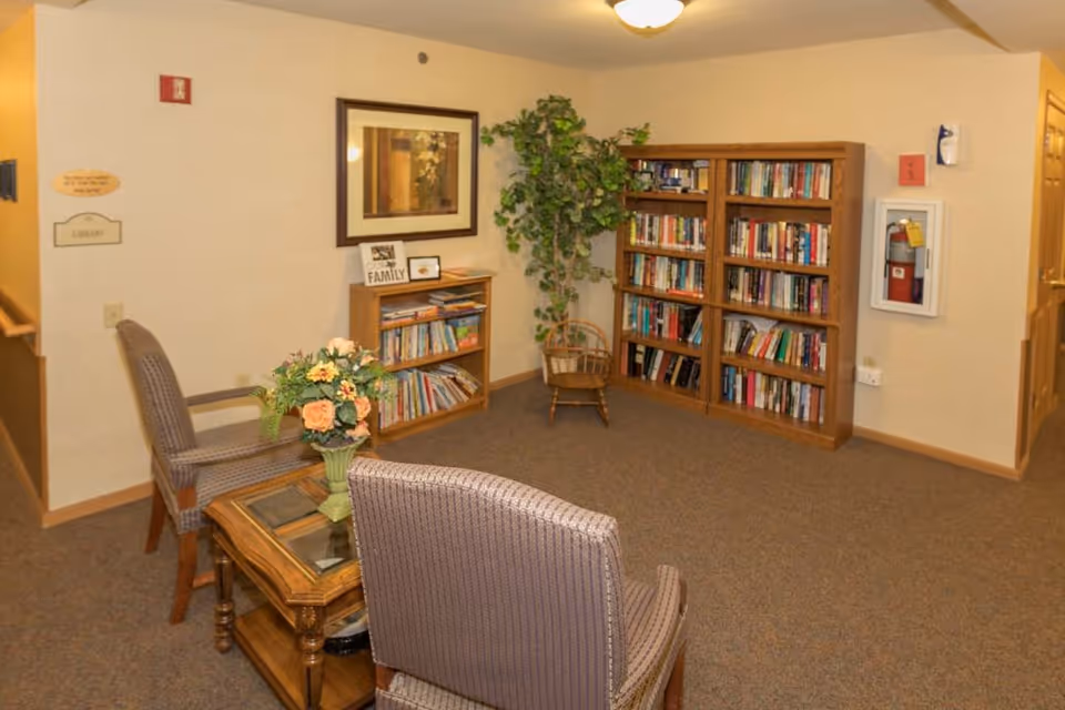 A cozy reading nook in an assisted living facility featuring two upholstered chairs around a wooden coffee table with a floral arrangement. There are two wooden bookshelves filled with books, a framed picture on the wall, a potted plant, and a small wooden chair. The walls are beige, and there is a fire extinguisher mounted on the wall.
