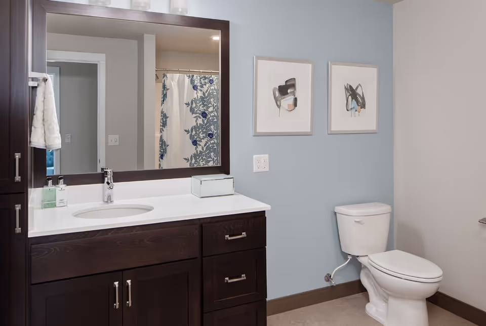 A clean and modern bathroom featuring a dark wood vanity with a white countertop and an oval sink. Above the vanity is a large mirror with a dark wood frame, reflecting a shower curtain with a blue floral pattern. To the right of the vanity is a white toilet. The walls are painted light blue and beige, adorned with two abstract art pieces in light-colored frames.