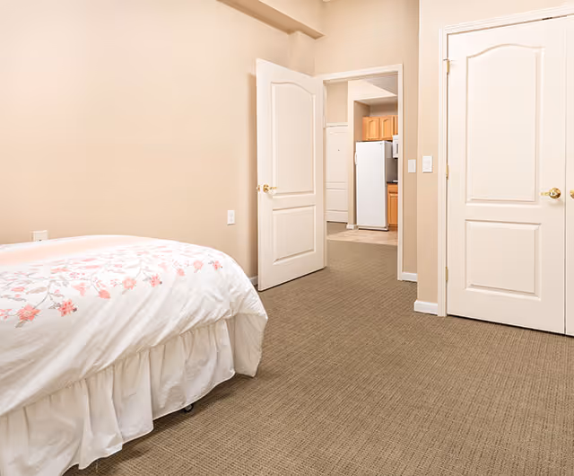 A bedroom with beige walls and carpet, featuring a bed with white and pink floral bedding on the left side. Two white doors are visible, one open showing a view into a kitchen area with wooden cabinets and a white refrigerator.