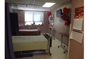 A room with two hospital-style beds, each with a different colored blanket, positioned side by side near a window with blinds. The room has light-colored walls decorated with balloons and pictures. There is a small table on wheels and a calendar hanging on the wall.