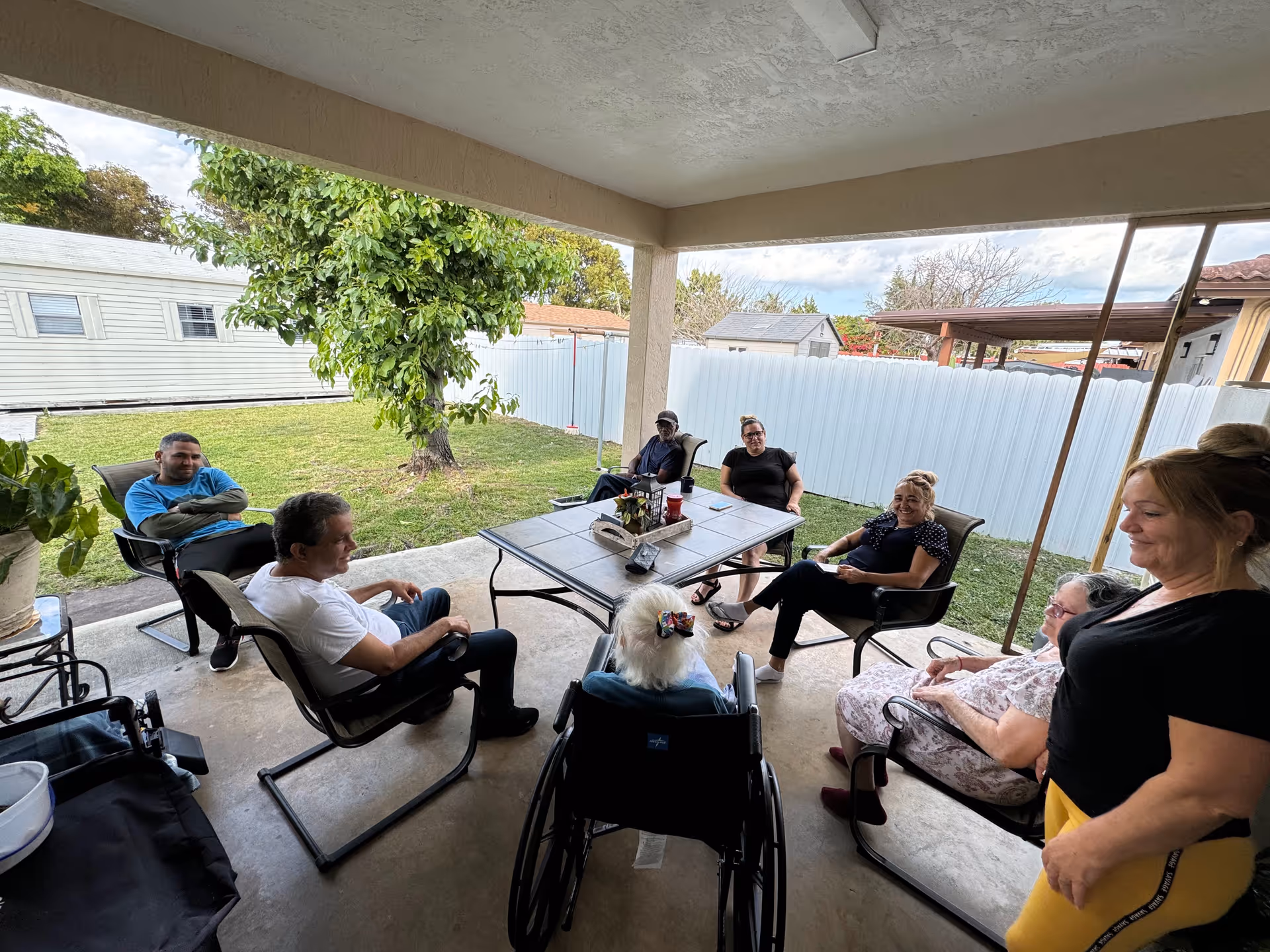 A group of people sitting and chatting around a rectangular table under a covered patio in a backyard. There are seven adults seated on chairs and one person in a wheelchair, with a green lawn, a tree, and a white fence in the background.