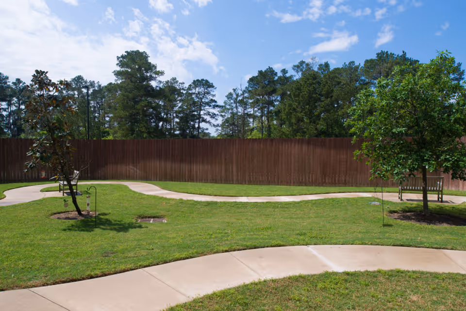 A grassy courtyard with winding concrete paths, small trees, benches, and a tall wooden fence with trees and blue sky beyond.