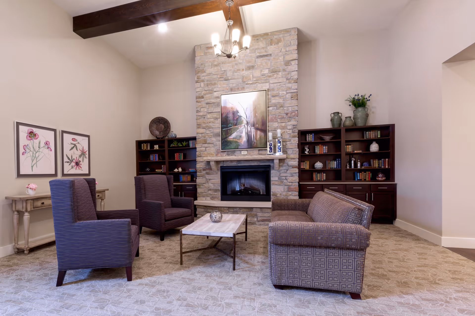 A cozy living room with a stone fireplace in the center, flanked by two dark wood bookshelves filled with books and decorative items. In front of the fireplace, there is a patterned sofa and two matching armchairs around a rectangular coffee table. The room has a beige carpet, light-colored walls, wooden ceiling beams, and a chandelier hanging from the ceiling. Two framed floral paintings hang on the left wall above a small wooden console table with a flower vase.