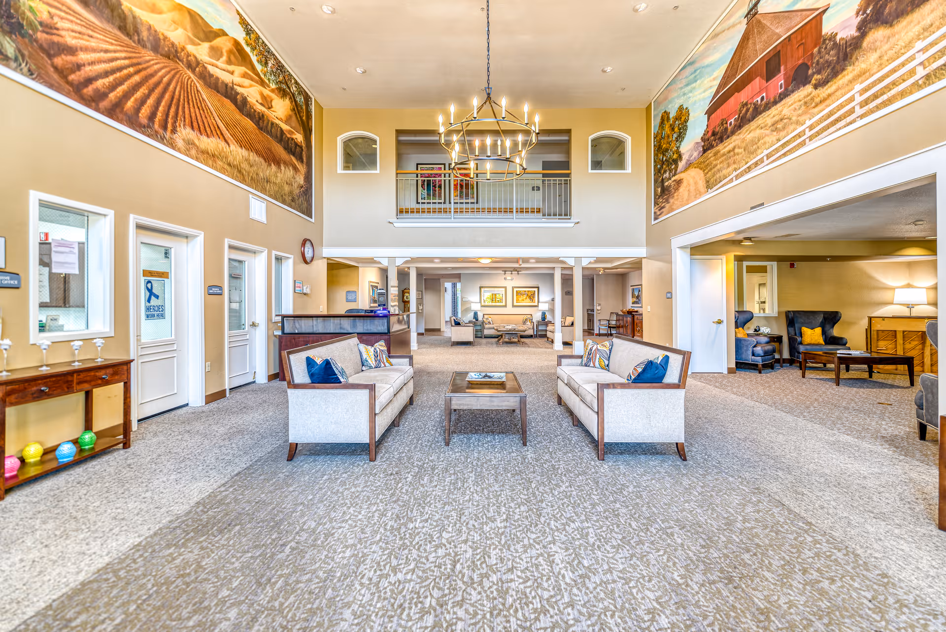 A spacious senior living facility common area with two beige sofas facing each other and a wooden coffee table in between. The room features high ceilings with a large chandelier, carpeted floors, and large murals of rural landscapes on the upper walls. There are additional seating areas with armchairs and tables visible in the background and to the right. Doors and windows line the left wall, and a balcony overlooks the space from the upper level.