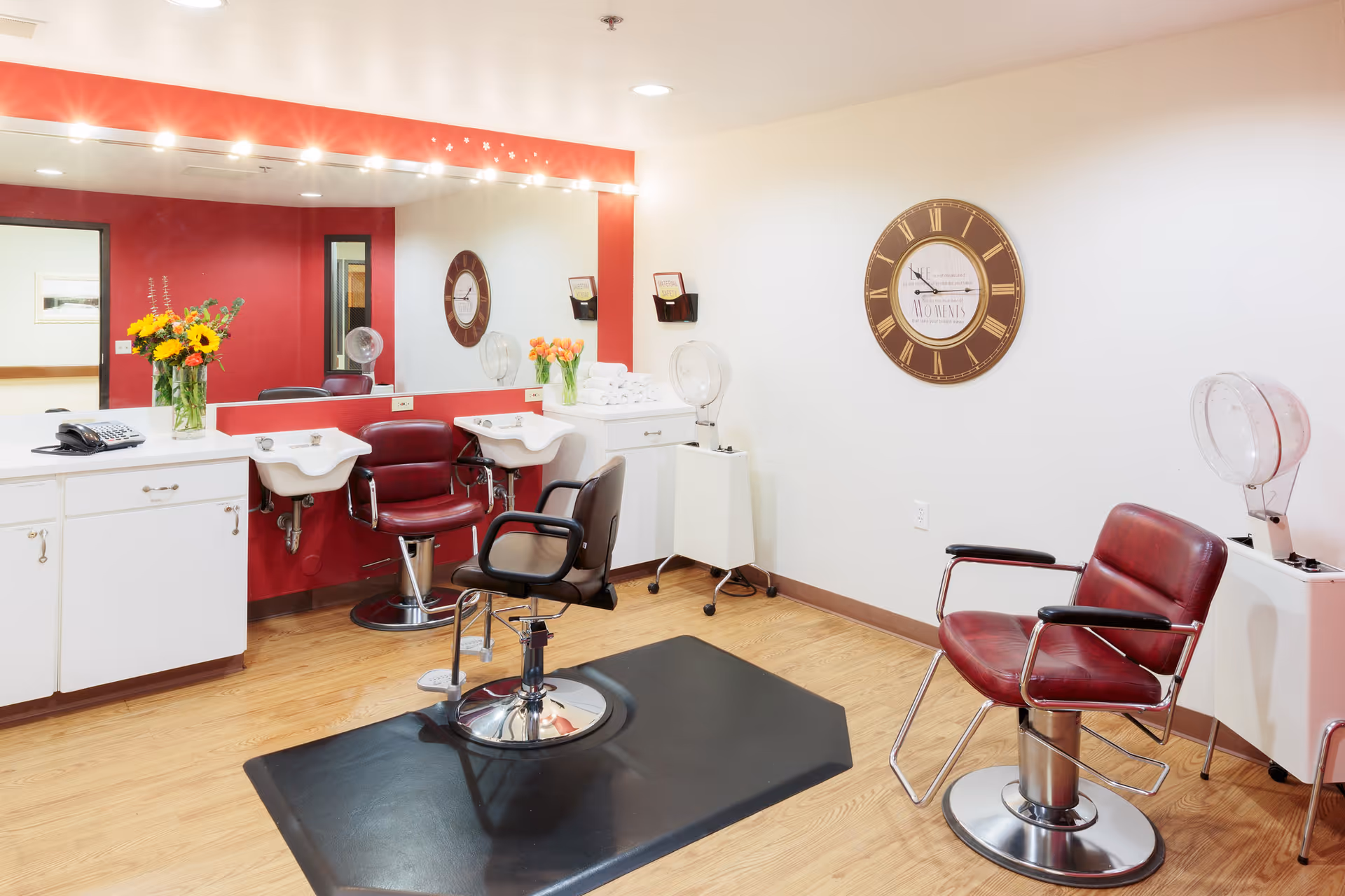 Interior of a hair salon area with red salon chairs, two white sinks, a large mirror with lights above it, a wall clock, and flower vases on white cabinets. The floor is wooden and the walls are painted white and red.
