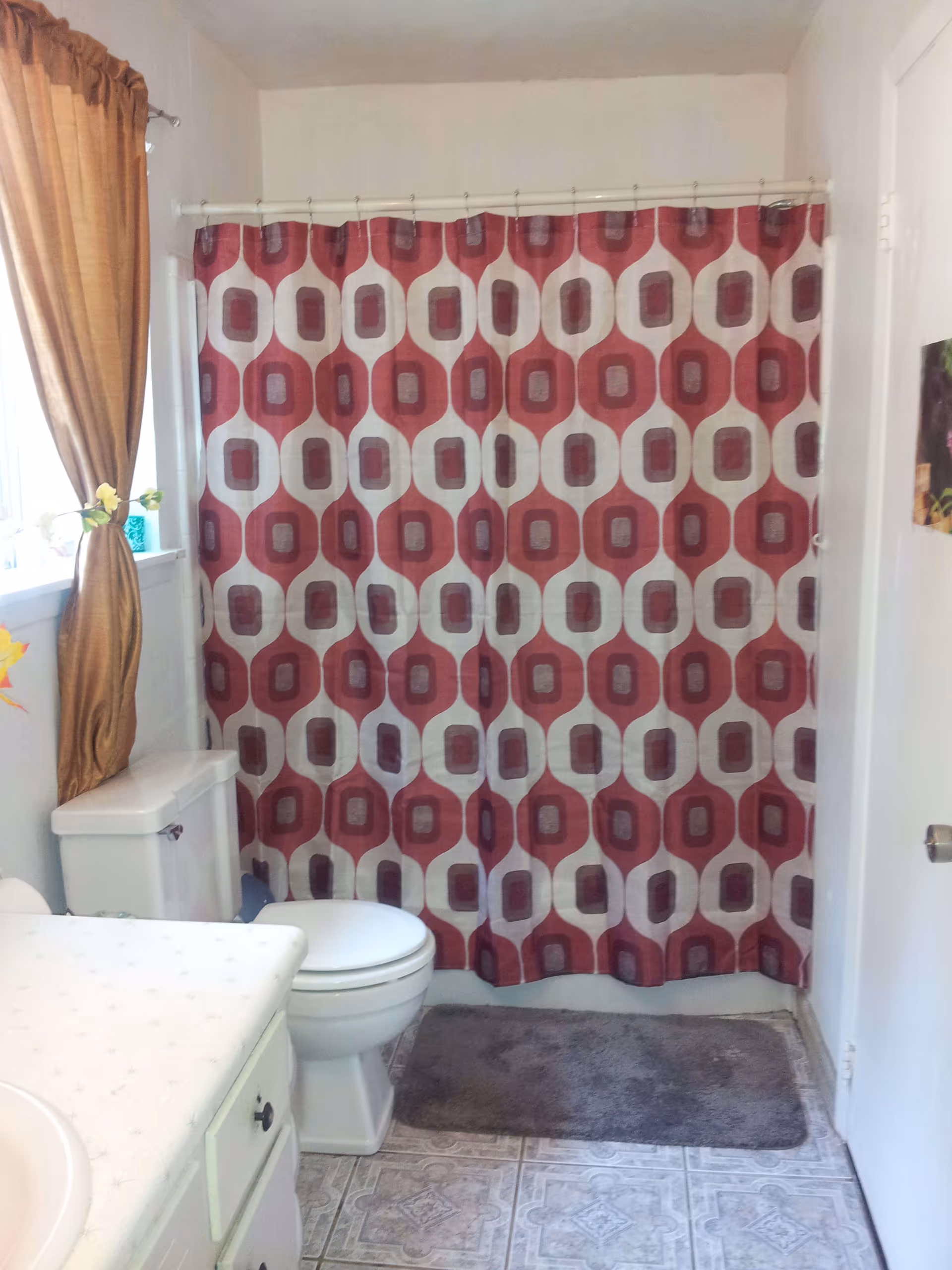 Bathroom with a white toilet, a countertop with a sink, a window with brown curtains, and a shower with a red and white patterned curtain. There is a gray bath mat on the tiled floor.