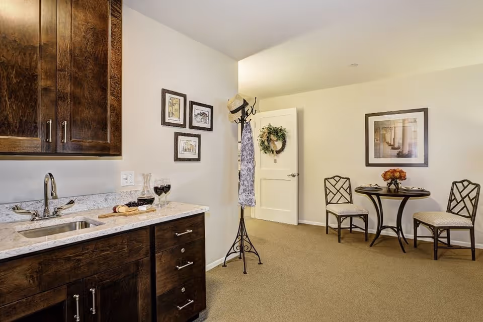 Interior view of a room with a small kitchenette on the left featuring a sink, countertop with a wine decanter, two glasses of red wine, and a cheese board. On the right side, there is a small round table set for two with a flower centerpiece and two chairs. The walls are decorated with framed pictures, and a coat rack with a hat and scarf stands near an open door adorned with a wreath.