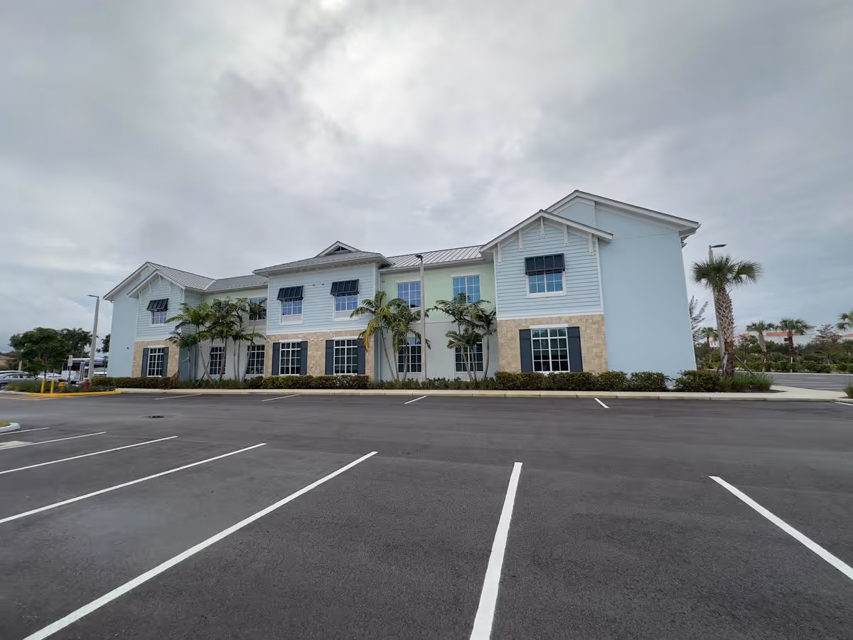 Two-story light blue building with palm trees in front and an empty parking lot under an overcast sky.