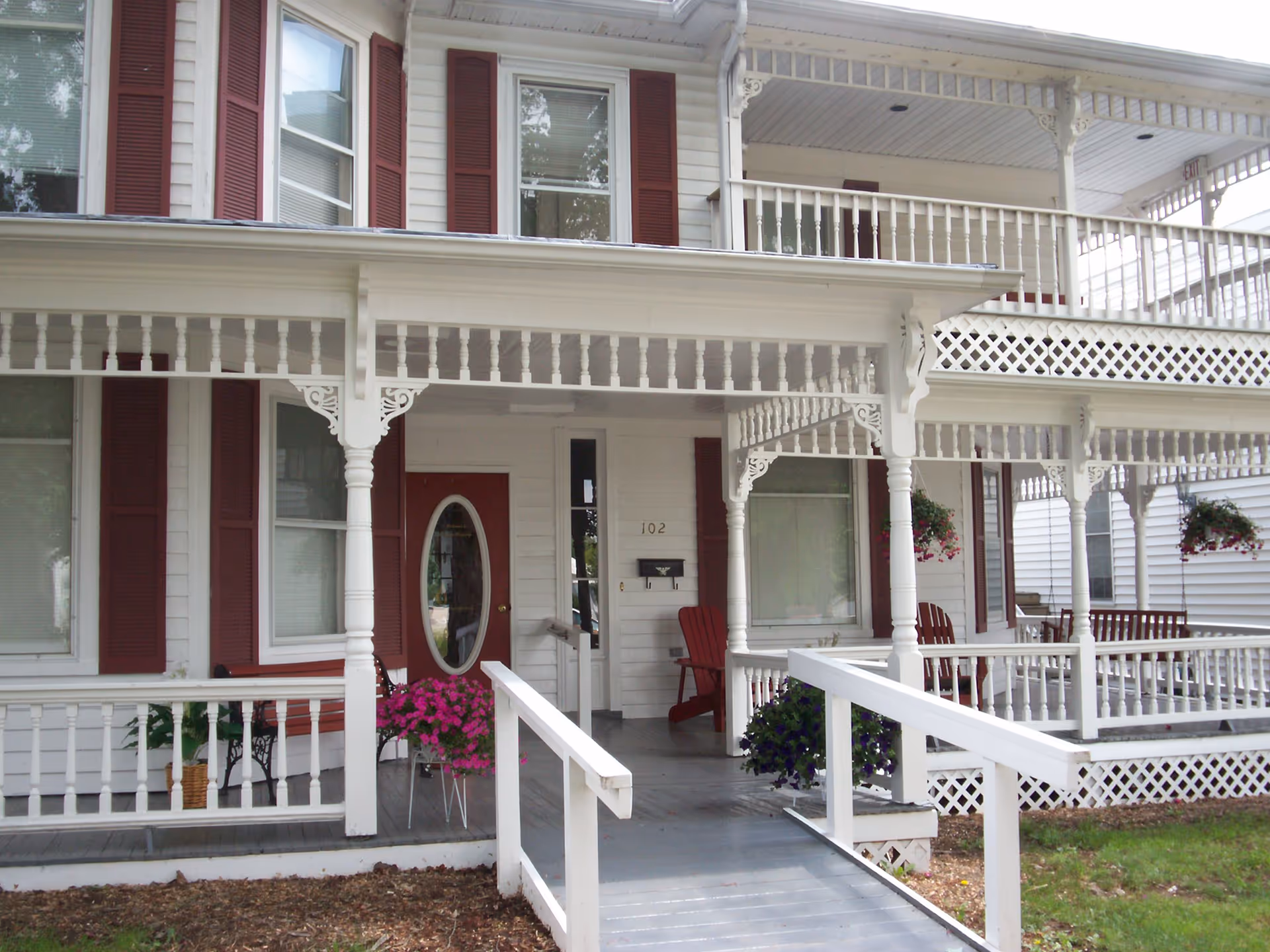 Two-story white building with ornate porch columns, red shutters, and hanging baskets.