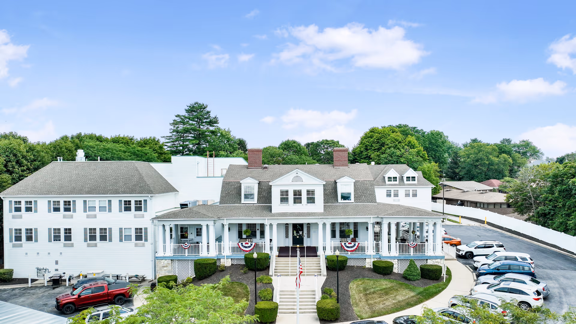 Front exterior view of The Terraces at Westminster facility, showing a large white building with a covered porch decorated with patriotic bunting. There are stairs leading up to the entrance, surrounded by neatly trimmed bushes and a curved driveway with parked cars. Trees and a blue sky with some clouds are visible in the background.
