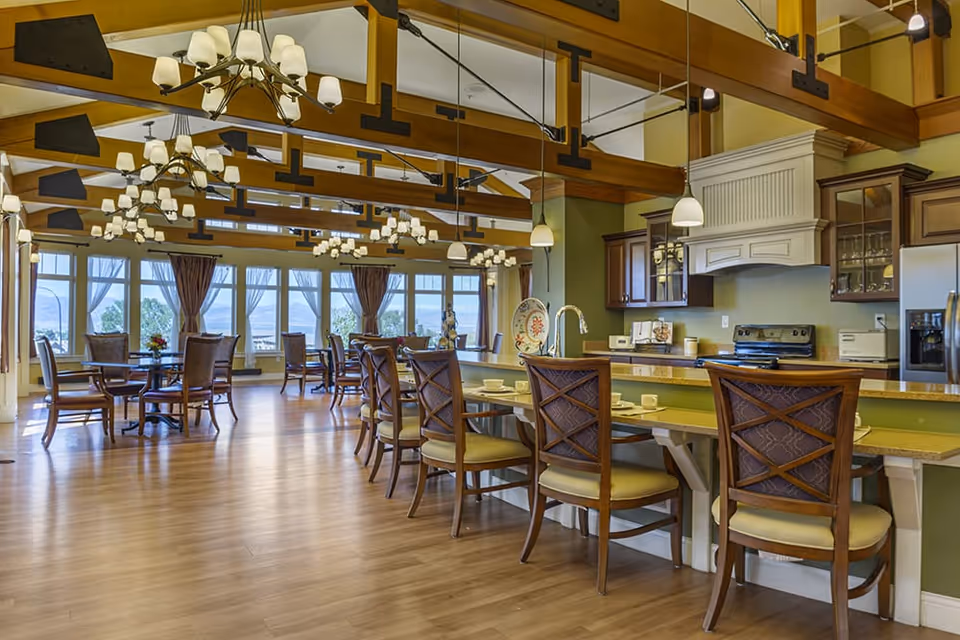 Bright communal dining room with exposed wood beams, multiple tables and a breakfast bar with chairs facing an open kitchen and large windows.
