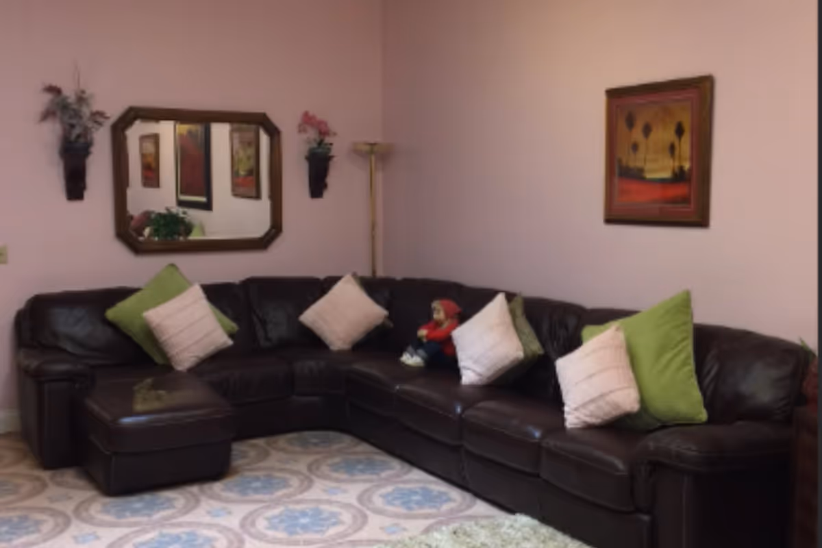 Dark brown leather sectional sofa with green and white pillows in a pink-walled living room with a mirror, wall art, and a floor lamp.