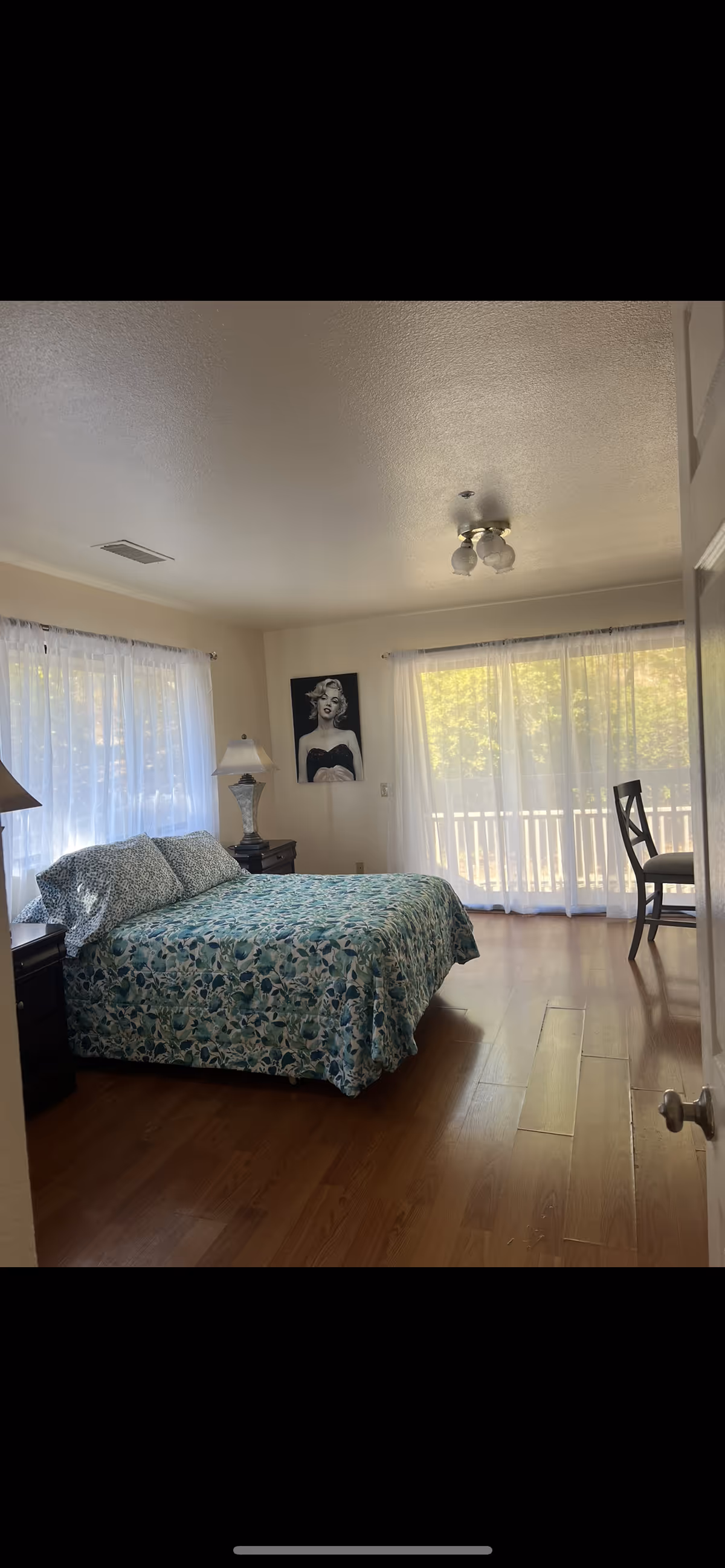 Bright bedroom with a patterned bed, nightstands and lamp, a Marilyn Monroe portrait, and large windows with sheer curtains leading to a balcony.