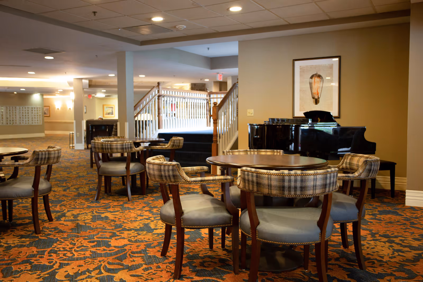 A spacious common area in an assisted living facility featuring round wooden tables with cushioned chairs upholstered in a plaid pattern. In the background, there is a black grand piano against a beige wall with a framed picture of a vintage light bulb above it. A staircase with white railings leads to an upper level, and the floor is covered with a patterned carpet in shades of orange and blue.