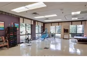 A spacious rehabilitation room with exercise equipment including a stationary bike and parallel bars. The room has large windows allowing natural light to enter, purple walls, and a shiny tiled floor.