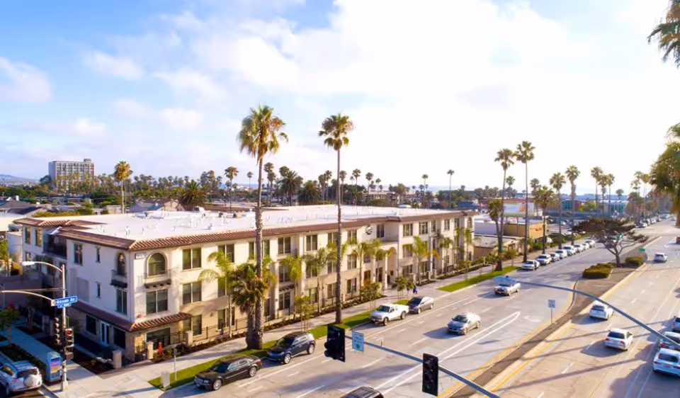Three-story Mediterranean-style building with palm trees along a busy coastal street and cars parked nearby.