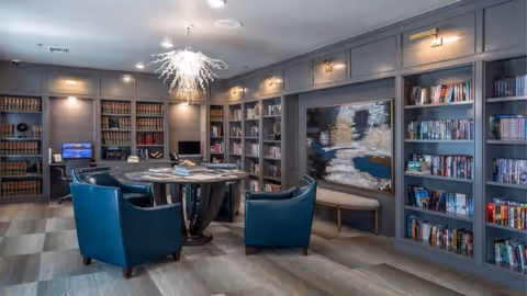 A cozy library or reading room with built-in bookshelves filled with books along the walls, a round table in the center surrounded by four teal armchairs, a modern chandelier hanging from the ceiling, and a large abstract painting on the wall.