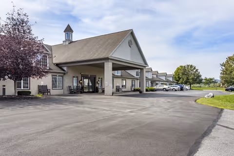 Exterior view of a senior living facility with a covered entrance, several parked cars, and a clear sky. The building has light-colored brick walls, multiple windows, and a small cupola on the roof. There are trees and green grass surrounding the paved driveway and parking area.