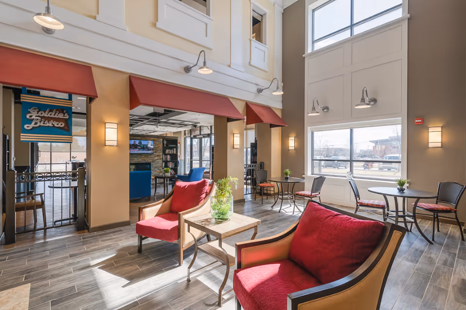 Bright and spacious common area with large windows letting in natural light. The room features comfortable seating with red cushioned chairs and small tables with plants. There is a sign for 'Goldie's Bistro' indicating a dining area nearby. The walls are painted beige with modern light fixtures and wood-look flooring.
