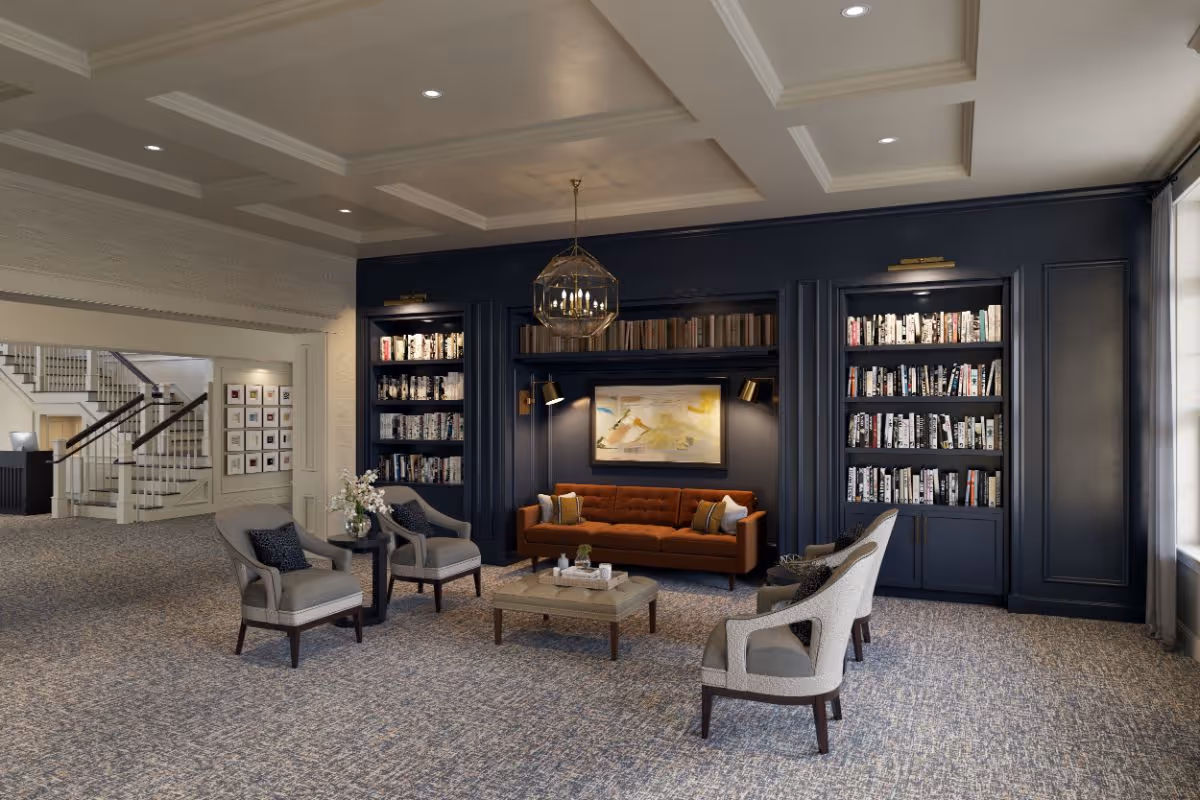 A cozy and elegant living room area with a brown sofa, four gray armchairs, a coffee table, and built-in dark blue bookshelves filled with books. The room features a decorative ceiling with recessed lighting, a large window with curtains, and a staircase visible in the background.