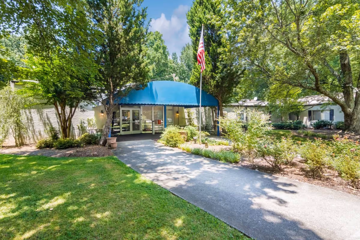 Walkway leading to the front entrance of a building with a blue awning, an American flag on a pole, and surrounding trees and landscaping.