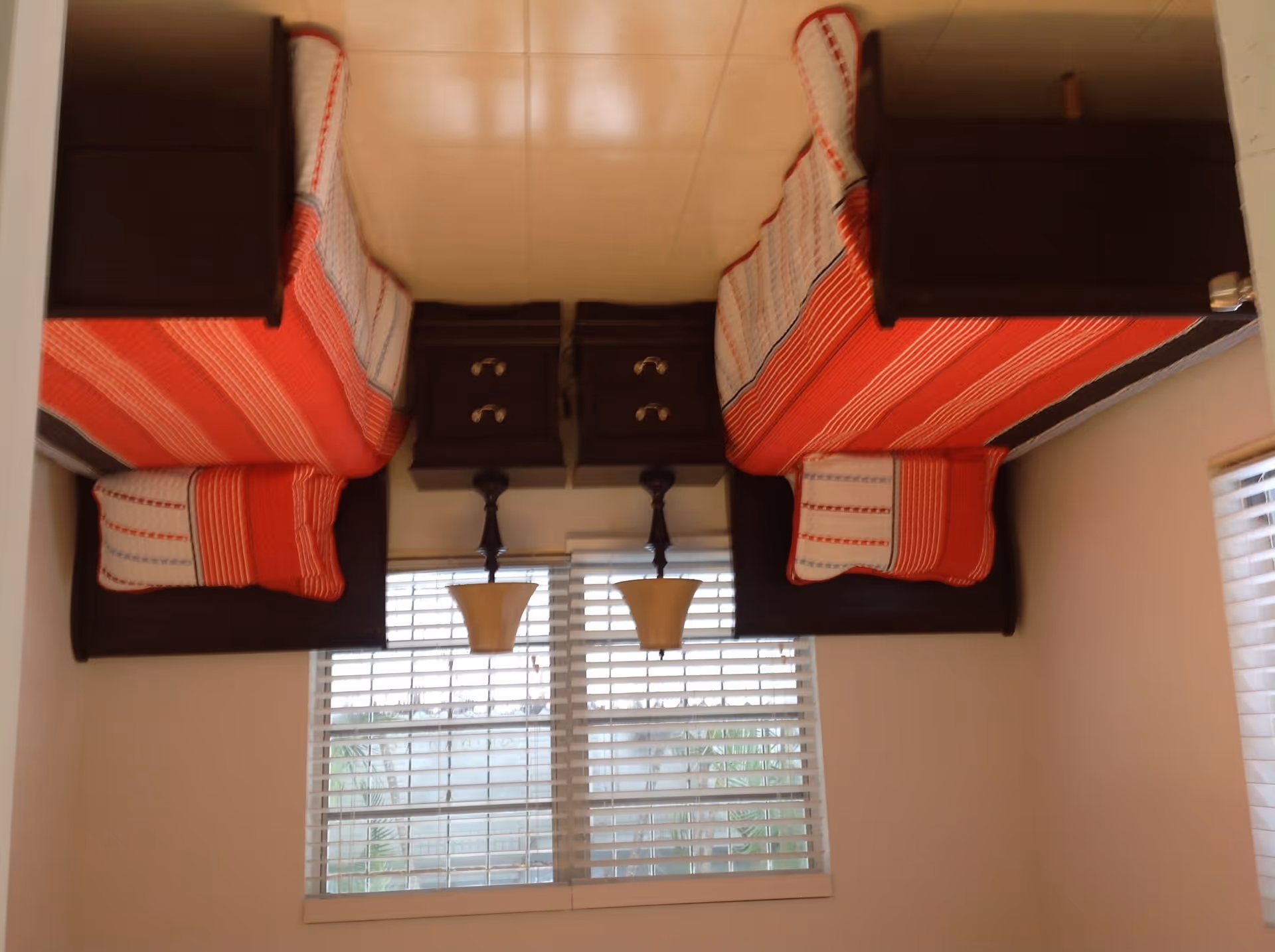 A bedroom with two twin beds placed parallel to each other, each with a red and white striped blanket and matching pillow. Between the beds is a dark wooden nightstand with two drawers, and a lamp with a beige lampshade on top. A window with white blinds is behind the nightstand, letting in natural light.