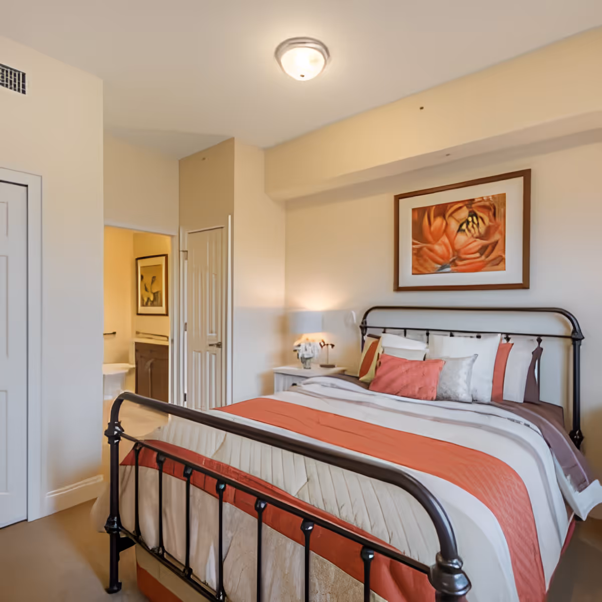 A cozy bedroom with a metal frame bed dressed in a striped orange, white, and beige comforter and multiple pillows. A nightstand with a lamp and a small plant is beside the bed. A framed floral artwork hangs on the wall above the bed. The room has beige walls and carpeted floor. An open door reveals a glimpse of an adjoining bathroom.