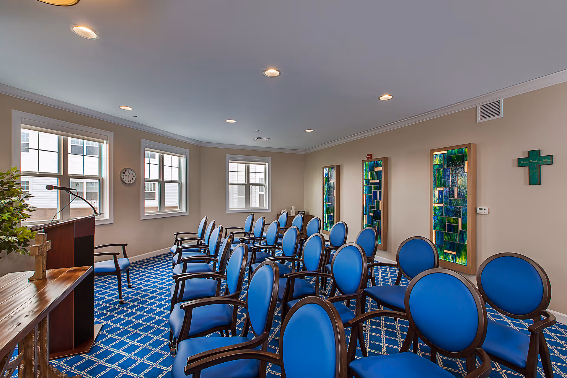 A small chapel or meeting room with rows of blue cushioned chairs arranged facing a wooden podium with a microphone. The room has three windows letting in natural light, a blue patterned carpet, beige walls, three vertical stained glass panels on one wall, and a green cross mounted on the wall.