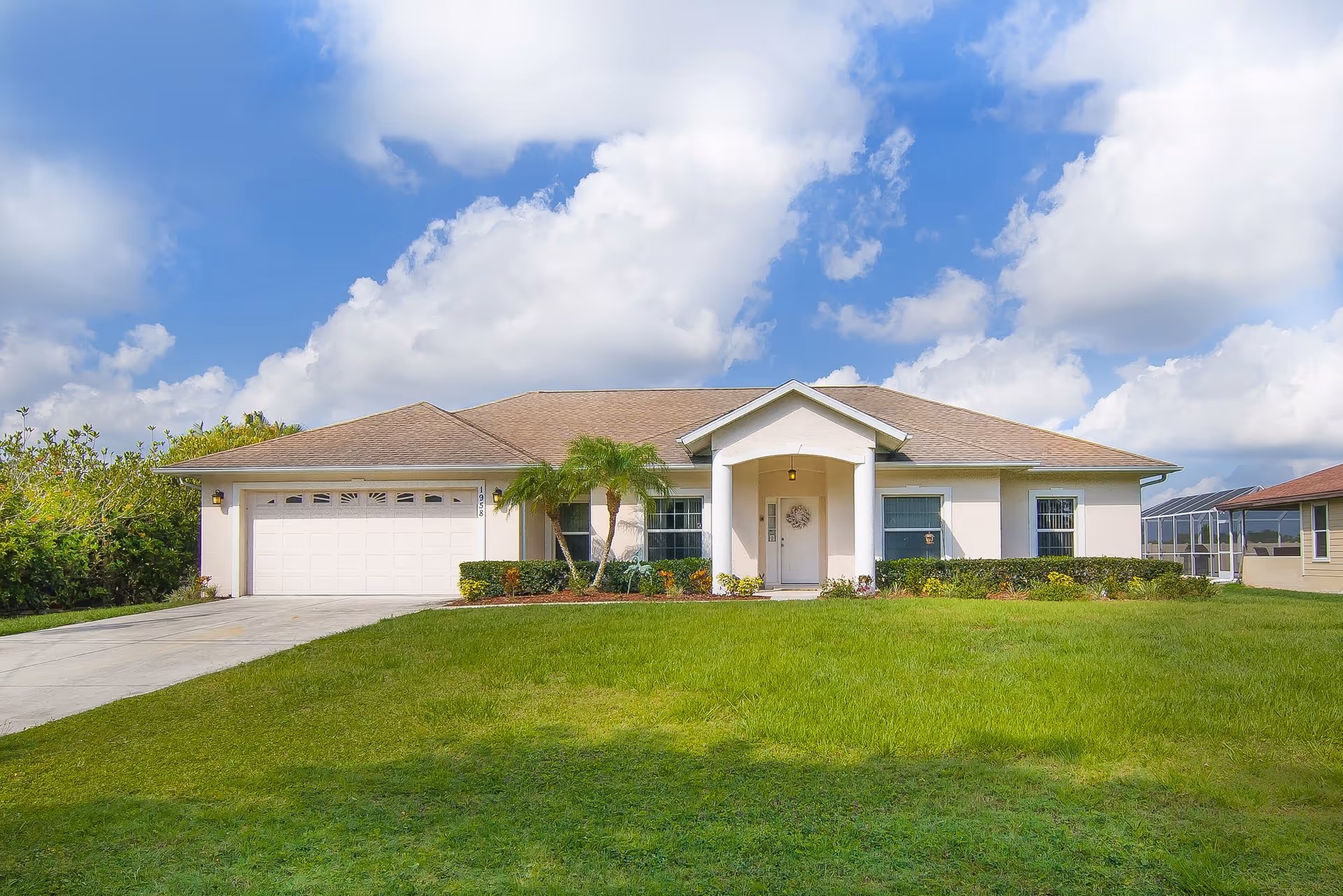 Single-story house with a beige exterior, a two-car garage, a front porch with two white columns, and a well-maintained green lawn under a partly cloudy blue sky.