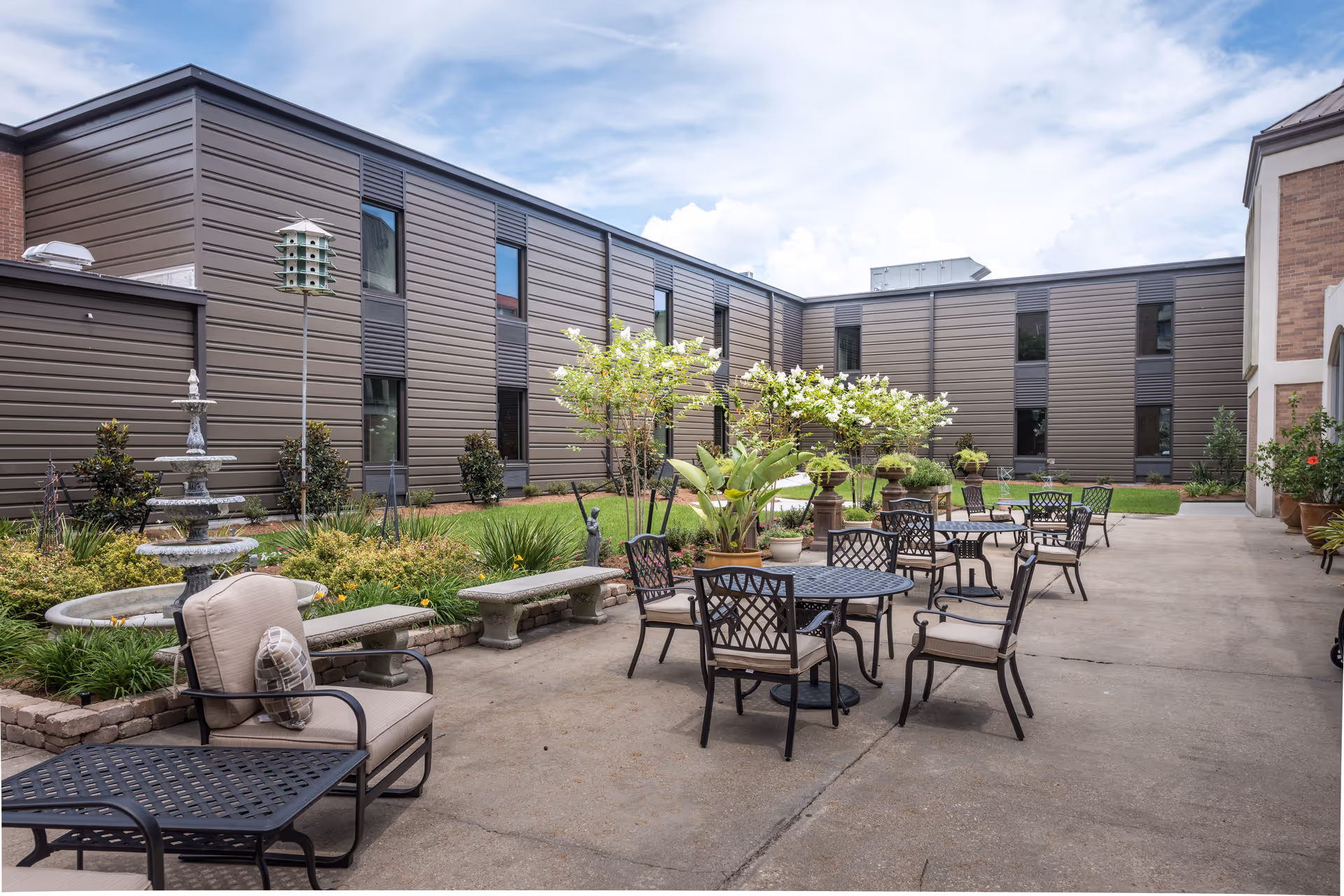 Landscaped courtyard with patio tables, chairs, benches, potted plants and a fountain beside a multi-story assisted living building.
