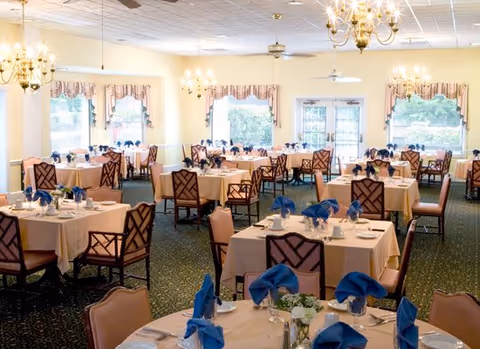 Large dining room with multiple set tables draped in light tablecloths and blue napkins, chandeliers, and windows overlooking greenery.