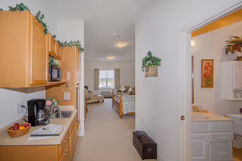 View of a senior living facility interior showing a kitchenette with wooden cabinets, a coffee maker, and a sink on the left. A hallway leads to a bedroom area with a bed and a window with curtains. On the right, there is a doorway leading to a bathroom with a sink, mirror, and floral decorations.