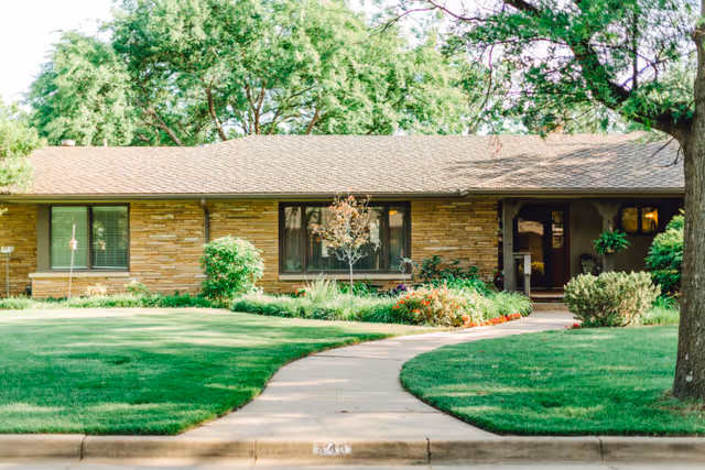 Front view of a single-story brick ranch-style home with a curved walkway, manicured lawn, and trees.