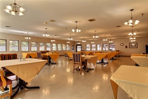 A spacious dining room with multiple tables covered in beige tablecloths and wooden chairs arranged around them. The room is well-lit with several ceiling light fixtures and large windows along one wall allowing natural light to enter. The floor is a light-colored polished surface, and the walls are decorated with a few framed pictures.