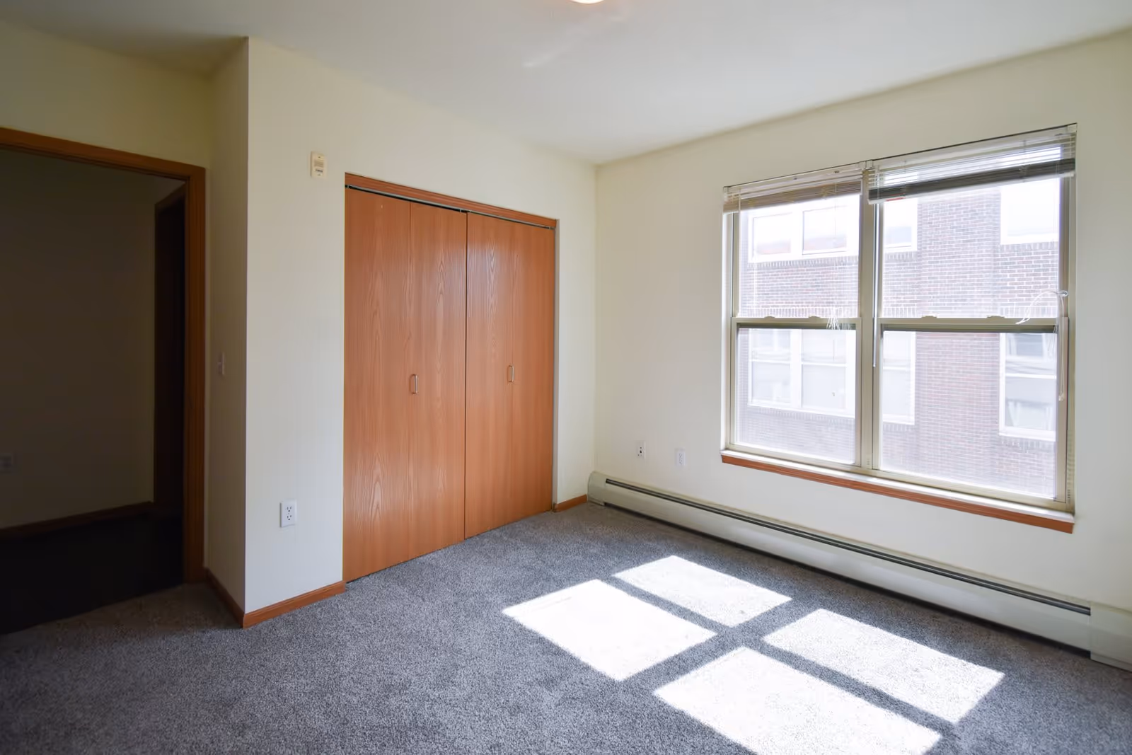Empty room in an apartment with beige walls, gray carpet, a wooden closet with double doors, and a large window letting in sunlight. The window shows a view of a brick building outside.