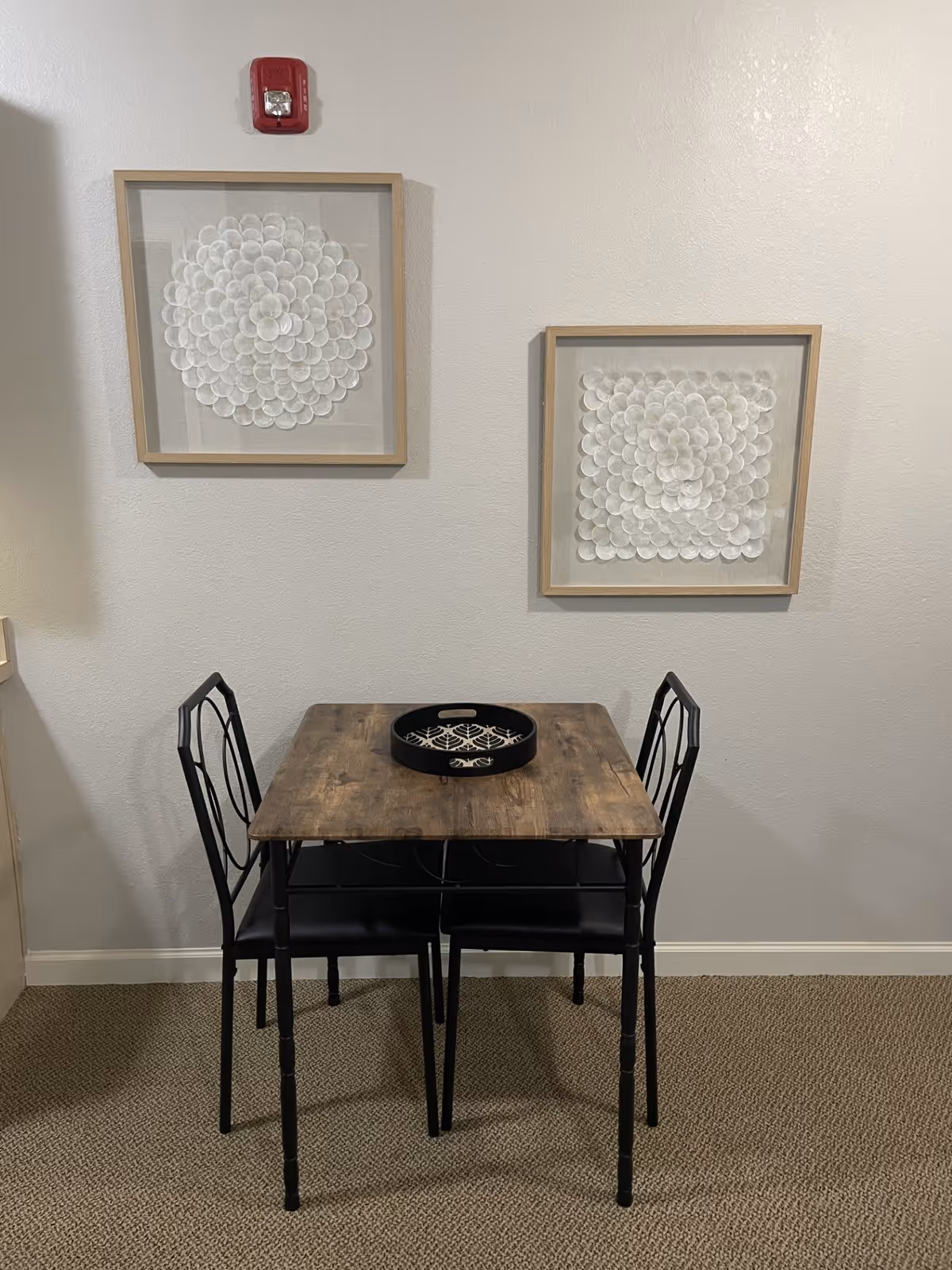 A small wooden table with a black decorative tray on top, accompanied by two black metal chairs with cushions, set against a plain white wall with two framed white floral artworks hanging above. A red fire alarm is mounted on the wall above the left artwork.